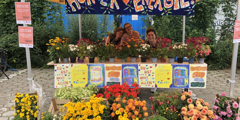 Ein bunter Marktstand unter einem gelben Pavillion mit Kisten voller Blumen im Vordergrund und auf einem Tisch sowie einem bunten Transparent auf dem