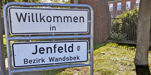 Ein zweigeteiltes Straßenschild am Wegesrand auf dem in schwarzer Schrift steht: Willkommen in Jenfeld, Bezirk Wandsbek