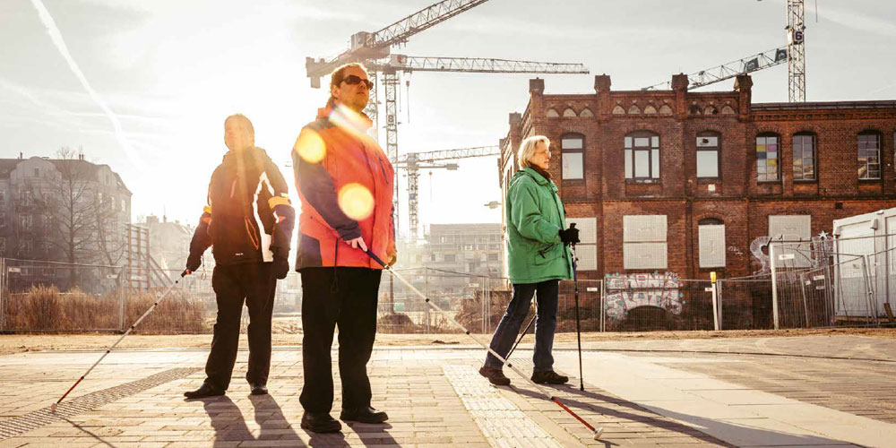 3 blinde Menschen gehen vorsichtig über einen Platz von Mitte Altona